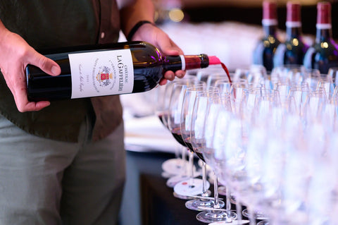 Person pouring red wine from a bottle labeled 'La Gafffeliere' into a row of wine glasses.