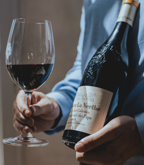 A person in a light blue shirt holds a bottle of red wine next to a filled wine glass. The bottle label reads 'Château La Nèrthé' and 'Château de Coudres'.