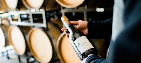 Person holding a bottle of wine in front of wine barrels